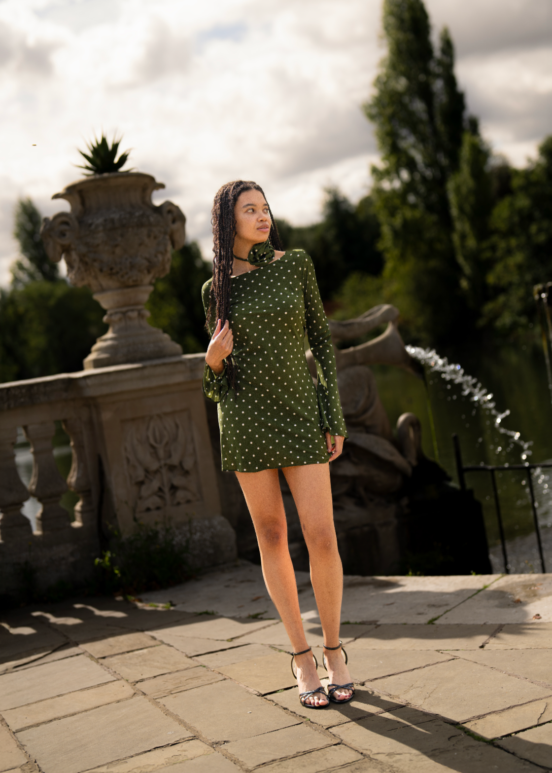Woman in a green polka dot dress standing outdoors with a fountain and garden in the background